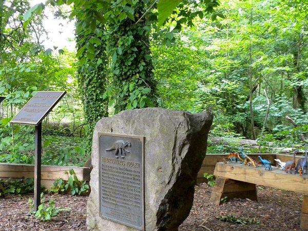 A stone marker with a bronze plaque and an interpretive sign at Hadrosaurus Park, trees behind.
