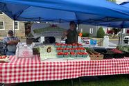A produce stand under a blue canopy at the Nazareth Farmers Market, with tomatoes on a red checked tablecloth