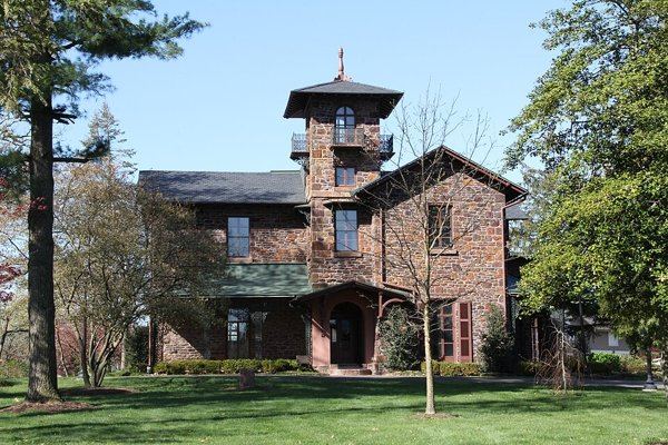 The front of Hollybush, a stone house with a tall square tower, an arched entry and a small balcony, behind a lawn and young trees.