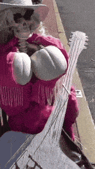 A skeleton scarecrow dressed as Dolly Parton, in sunglasses, a white cowboy hat and a pink fringe jacket, holding a white guitar on a sunny sidewalk.