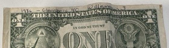 The top edge of a worn one-dollar bill with 1986 - Glassboro 40 Williamstown 3 handwritten across it in ink.