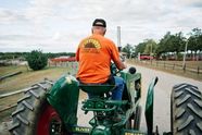 A farmer seen from behind drives a vintage tractor along a farm lane, with fields and trees behind.