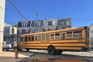 A yellow school bus turning a tight cobbled corner in Manayunk, a neighbor standing by its front